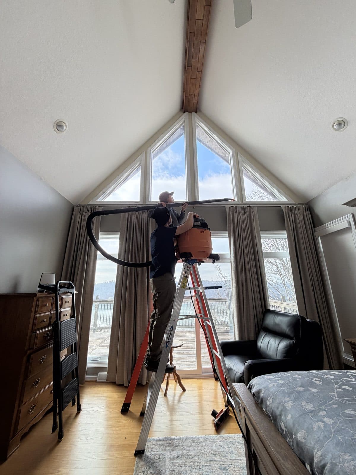 A Micro Cleaning Pro technician on a ladder, HEPA-vacuuming a window frame near the ceiling of a client's home.